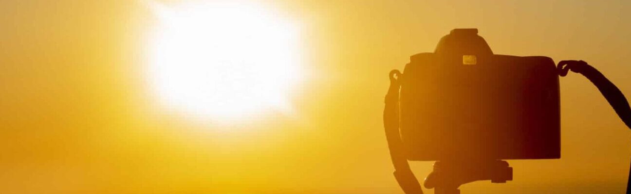 Silhouette of digital camera on a tripod in front of setting sun with orange sky.