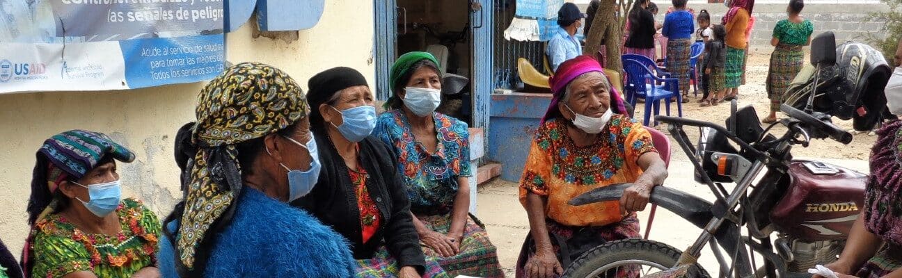 5 women wearing colourful clothing and face masks sit on a bench talking.