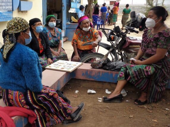 Five women wearing colourful clothing and face masks sit on a bench talking.
