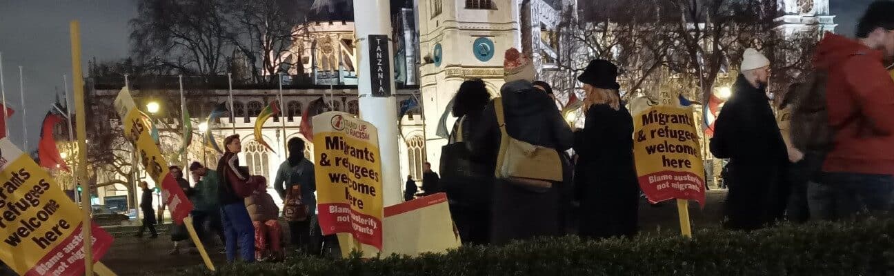 Photo outside the houses of Parliament of people standing around, placards that say "Migrants and refugees welcome here" and a handwritten cardboard sign leaning against a bush that says "Refugees welcome"