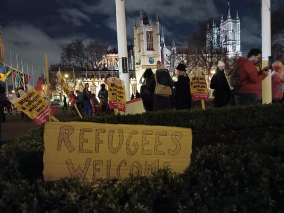 Photo outside the houses of Parliament of people standing around, placards that say "Migrants and refugees welcome here" and a handwritten cardboard sign leaning against a bush that says "Refugees welcome"