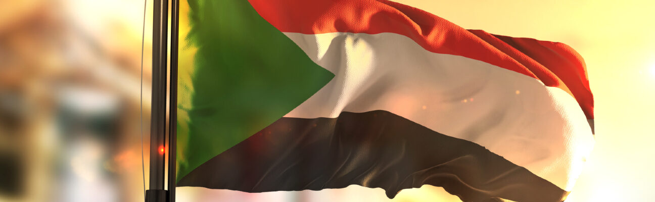 Sudan Flag Against City Blurred Background At Sunrise Backlight