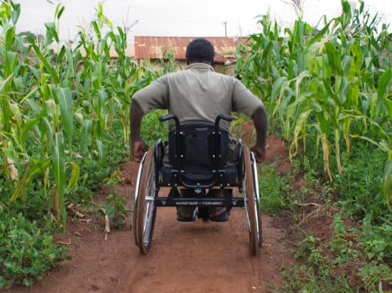 Man in a wheelchair pushes himself through a field towards a house.
