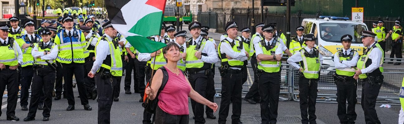 A lone woman stands before a wall of police. She holds high a large Palestine flag and waves it gently. Credit: Alisdare Hickson