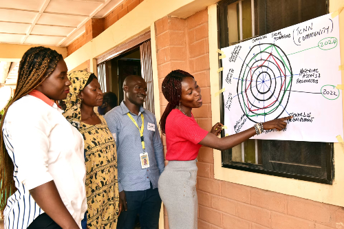 A group of Nigerian men and women crowd around a large piece of paper on a wall. One man writes on the paper as the group add their ideas relating to the "Personal relationships" spoke of the Light Wheel. Credit: Nanmet Anthony/Tearfund